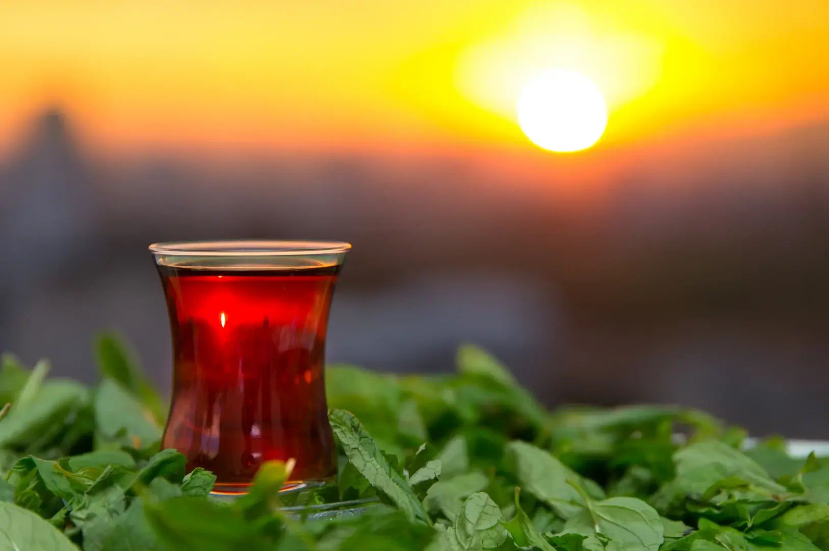 Yeşil yaprakların üzerinde bir çay bardağı, arka planda gün batımı manzarası.