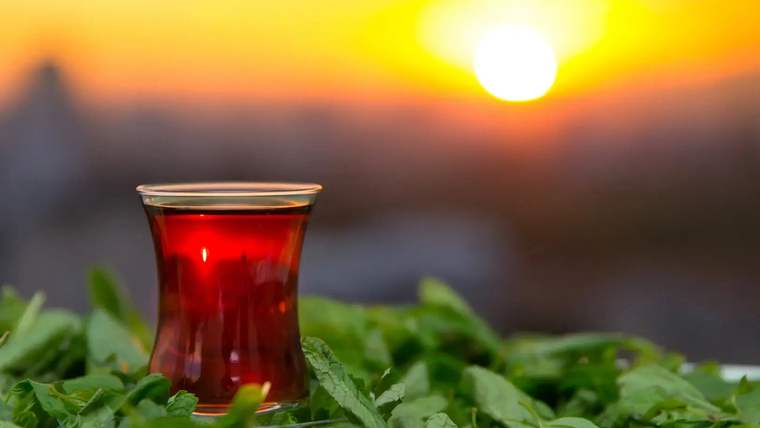 Yeşil yaprakların üzerinde bir çay bardağı, arka planda gün batımı manzarası.