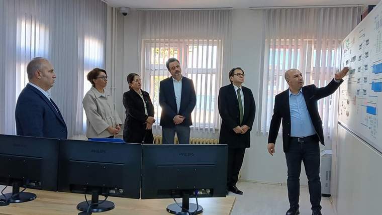 Bir grup insan, bir odada duvarda bulunan bir panoya bakıyor. Ön planda bir kişi, panoda bir şeyler gösteriyor.
