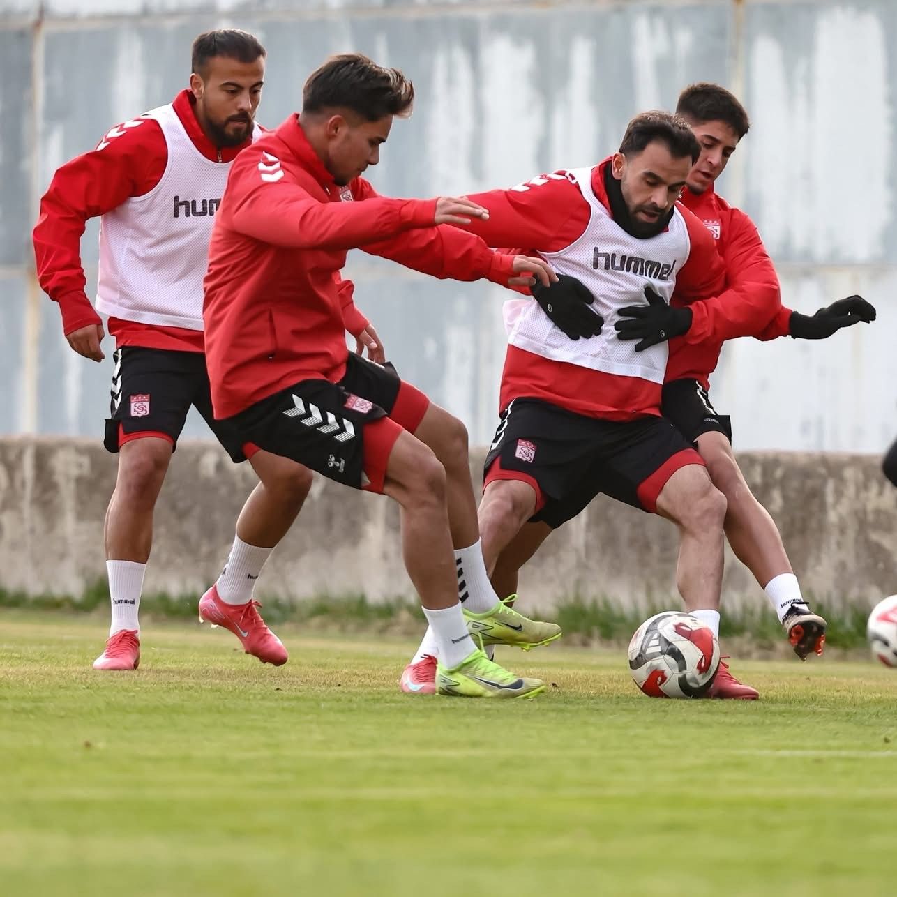 Kırmızı-beyaz formalar giyen dört futbolcu, çim sahada top için mücadele ederken görülüyor.