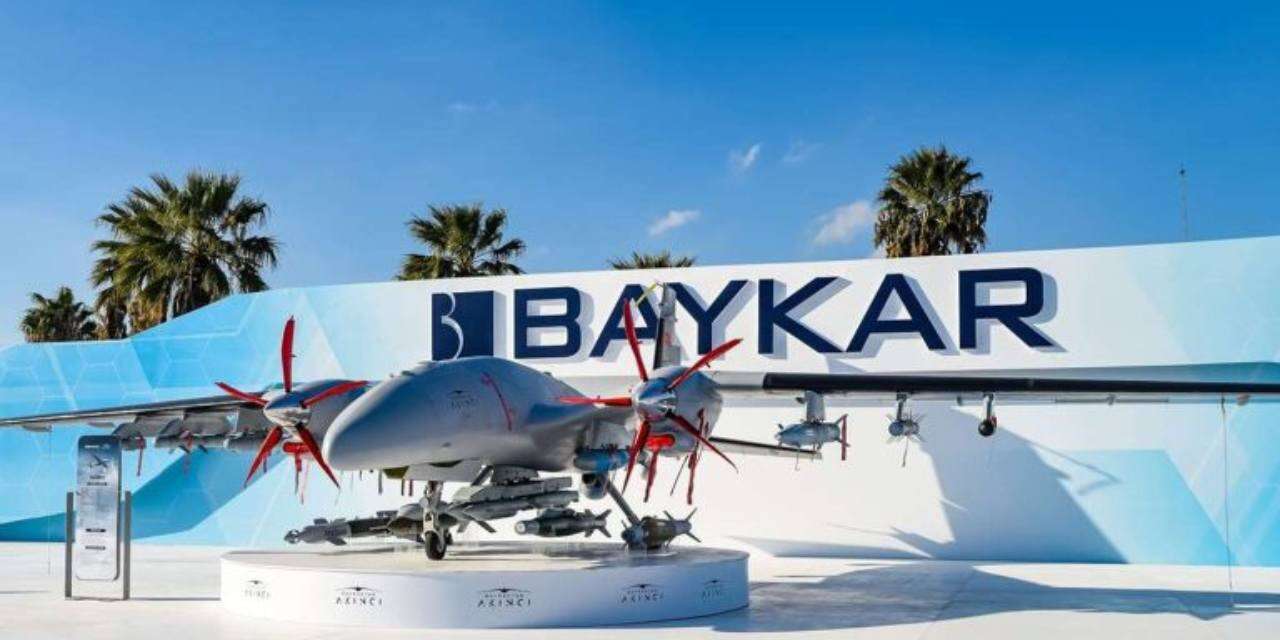 BAYKAR yazılı bir binanın önünde sergilenen drone, beyaz platform üzerinde duruyor; arkada palmiye ağaçları görülüyor.