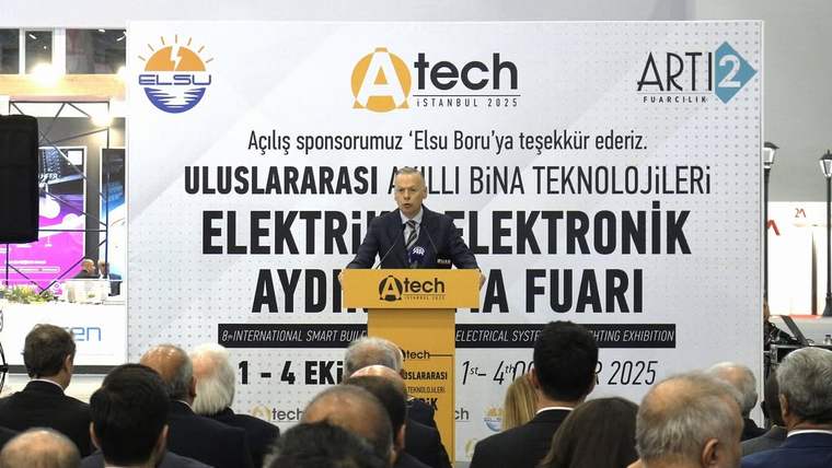 Kürsüde konuşan bir kişi, arkasındaki fuar pankartının önünde oturan kalabalığa sesleniyor.