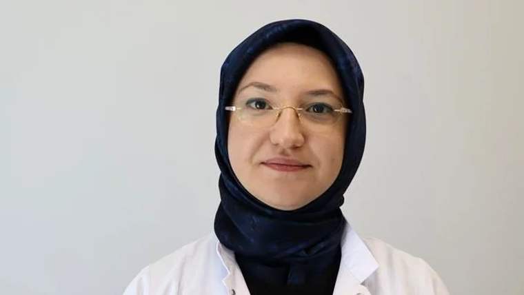 Beyaz önlük giymiş, başında mavi bir başörtüsü olan bir kadın, kameraya gülümsüyor.