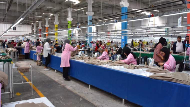 Geniş bir tekstil atölyesinde çok sayıda kişi, kumaş ve dikiş makinelerinin bulunduğu masalarda çalışıyor.