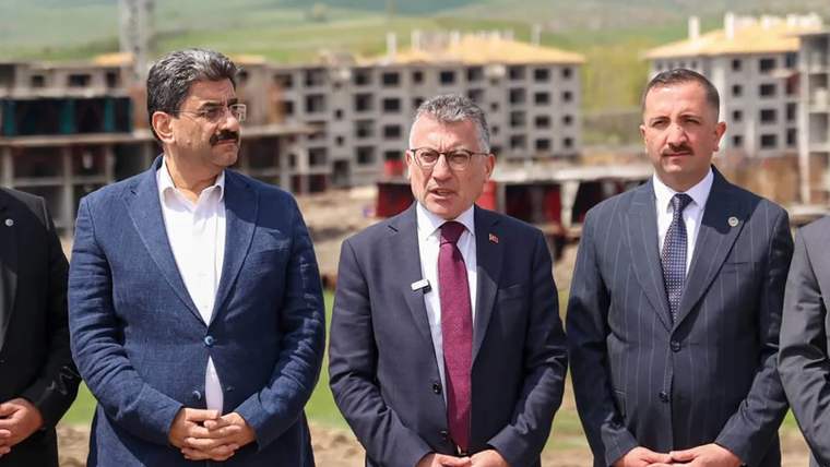 Üç kişi, inşaat alanında durarak kameraya bakıyor. Arkada tamamlanmamış binalar görünmekte.
