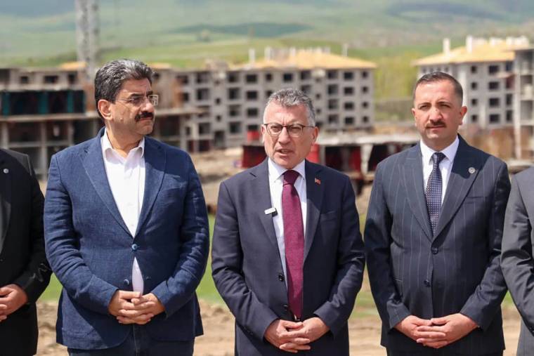 Üç kişi, inşaat alanında durarak kameraya bakıyor. Arkada tamamlanmamış binalar görünmekte.