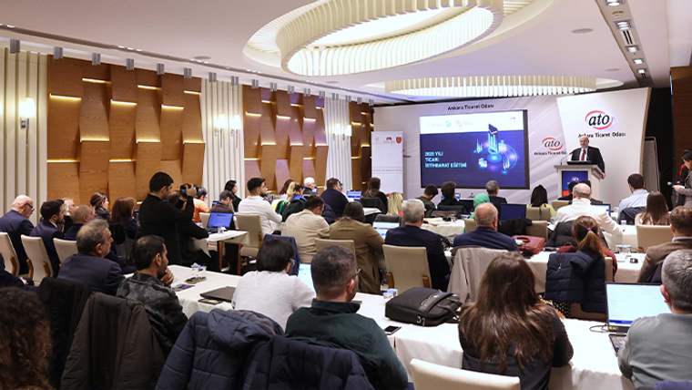 Toplantı salonunda sıralara oturan katılımcılar, ekrandaki sunumu ve kürsüde konuşan kişiyi izliyor.