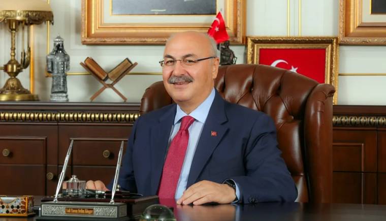Makam odasında takım elbiseli, gözlüklü bir kişi masasında oturuyor; arkasında Türk bayrakları ve tablolar bulunuyor.