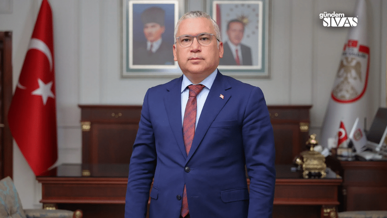 Mavi takım elbise giymiş bir kişi, arkasında Türk bayrağı ve portreler ile poz veriyor.