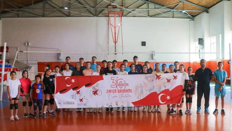 Kapalı spor salonunda voleybolcular, üzerinde Türk bayrakları ve Zafer Bayramı yazısı bulunan pankartı tutuyor.