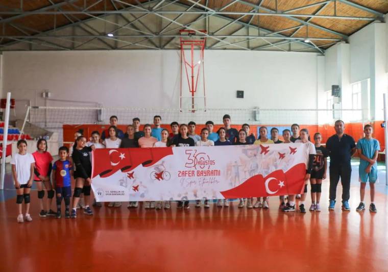 Kapalı spor salonunda voleybolcular, üzerinde Türk bayrakları ve Zafer Bayramı yazısı bulunan pankartı tutuyor.