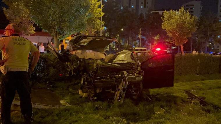 Gece aydınlatmasında ağır hasarlı bir otomobil, çim alanda dururken yanında kırmızı ışıklı araç bulunuyor.
