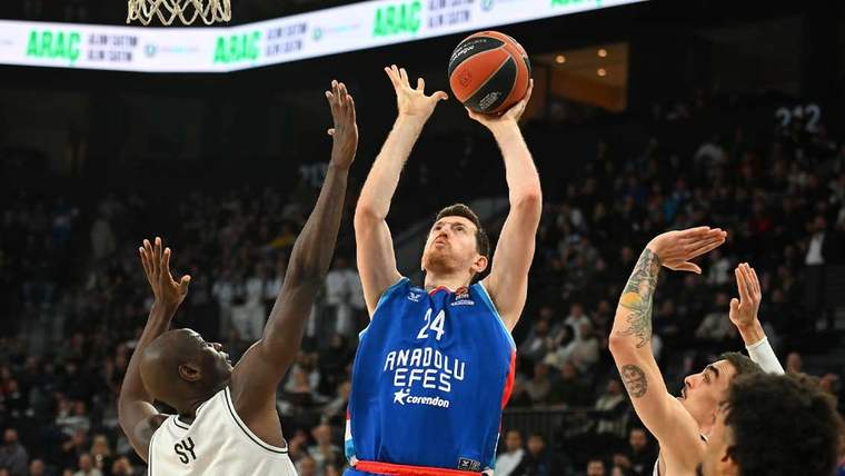 Anadolu Efes forması giymiş bir basketbolcu, topu potaya atmaya çalışıyor. İki rakip oyuncu savunma yapıyor.