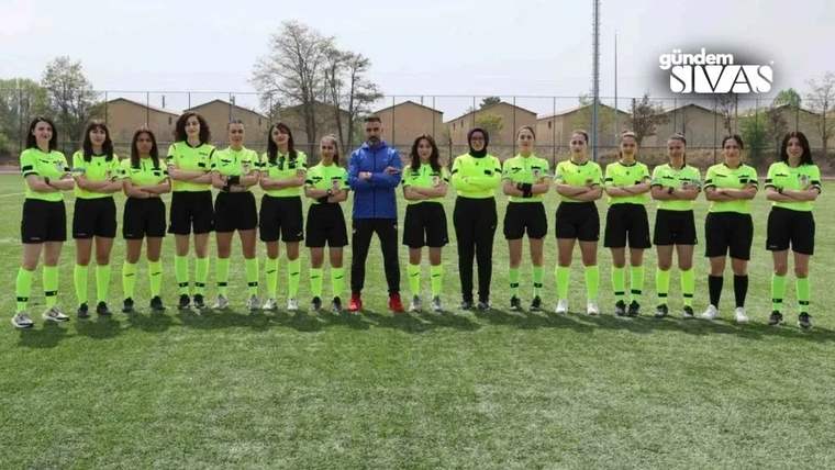 Futbol sahasında yeşil formalar giyen kadın hakemler, mavi giyimli bir kişiyle yan yana duruyor.