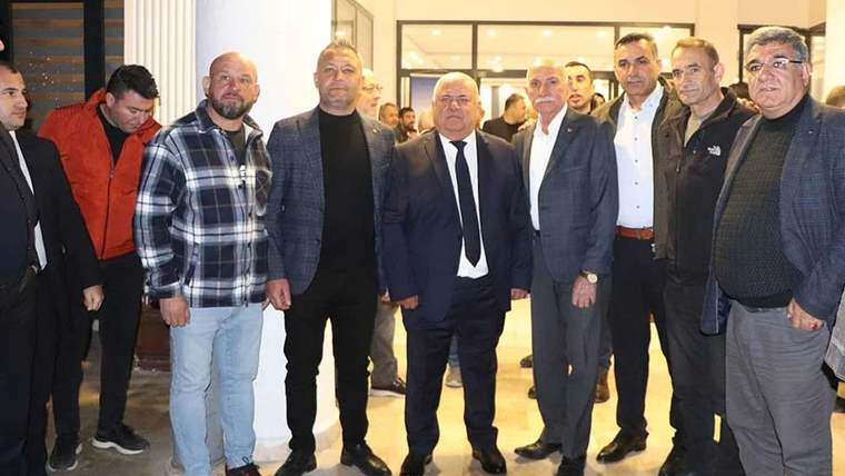 Kapalı bir salonda, takım elbiseli ve günlük giyimli bir grup erkek yan yana duruyor.