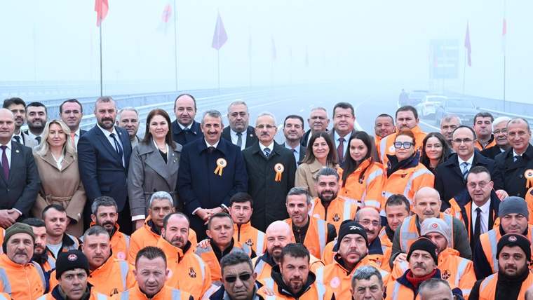 Açılış töreninde yer alan kalabalık, ön planda turuncu yelek giymiş çalışanlar ve arka planda bayraklar ile bir yol görünmektedir.
