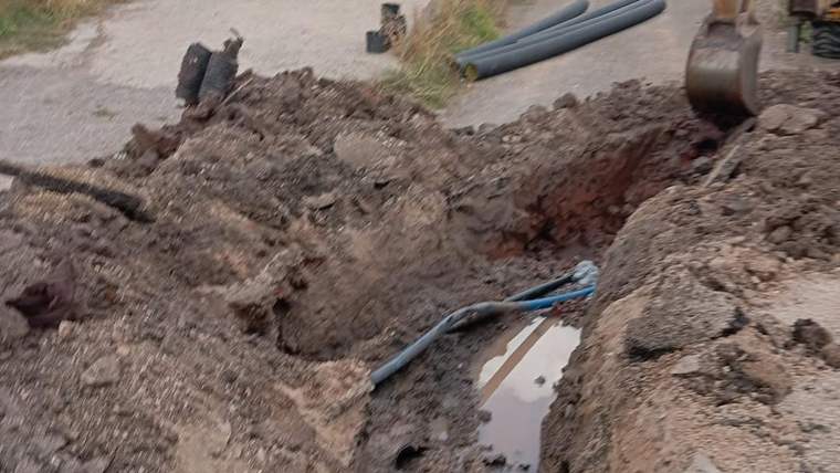 Toprak yolda açılmış kanalda mavi borular ve su birikintisi, yanında iş makinesinin kepçesi görülüyor.