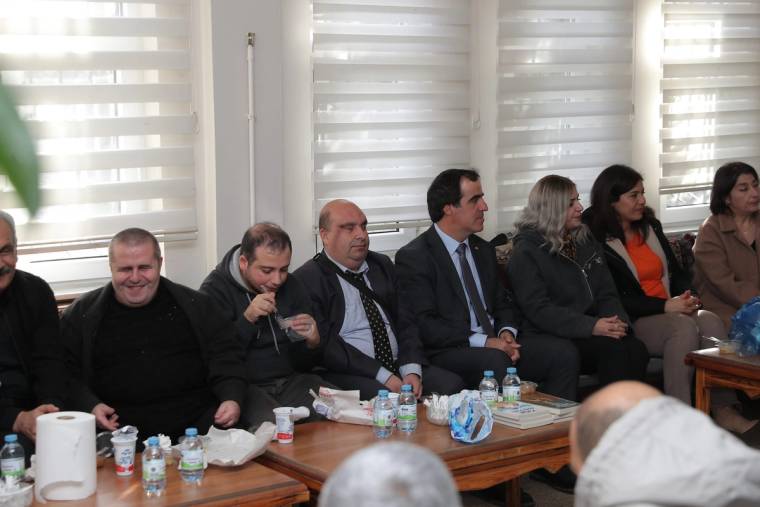 Mikrofon tutan bir kişi ve yanındaki kişiler, masada su şişeleri bulunan salonda oturuyor.