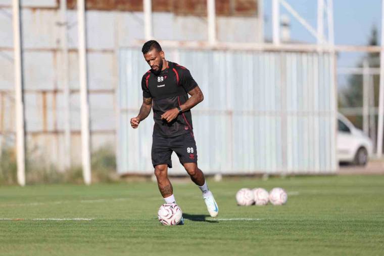 Futbol antrenman sahasında siyah forma giyen bir oyuncu, top sürerken görülüyor; çevrede futbol topları bulunuyor.