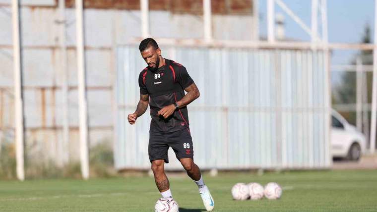 Futbol antrenman sahasında siyah forma giyen bir oyuncu, top sürerken görülüyor; çevrede futbol topları bulunuyor.