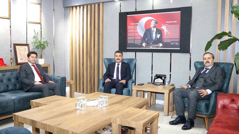 Bir ofiste üç erkek koltuklarda oturuyor; arkalarındaki ekranda takım elbiseli bir kişinin portresi görülüyor.