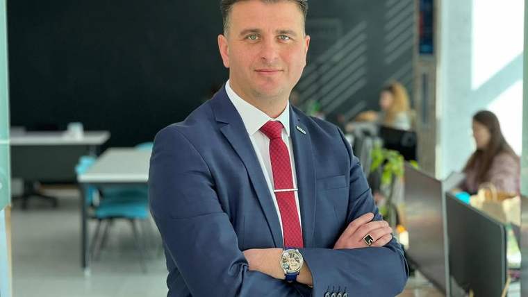 Lacivert takım elbiseli bir erkek, kollarını bağlayarak iç mekânda kameraya bakıyor.
