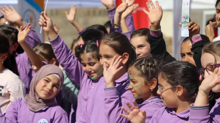 Mor ve pembe kıyafetli çocuklar, ellerini kaldırarak kalabalık içinde birlikte duruyor.