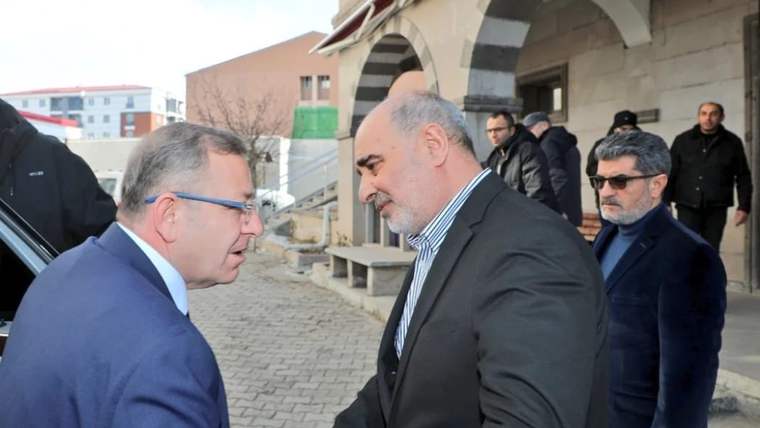 İki erkek dışarıda el sıkışıyor; arkalarında kemerli taş yapı önünde birkaç kişi bekliyor.