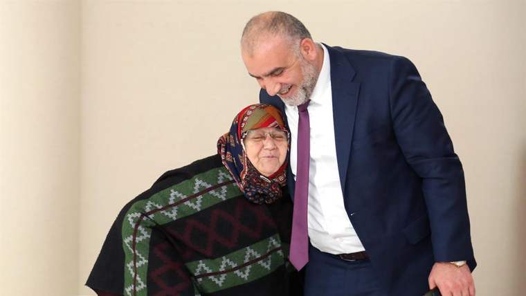 Takım elbiseli bir erkek, desenli başörtüsü ve şal takan yaşlı bir kadının yanında eğilerek duruyor.