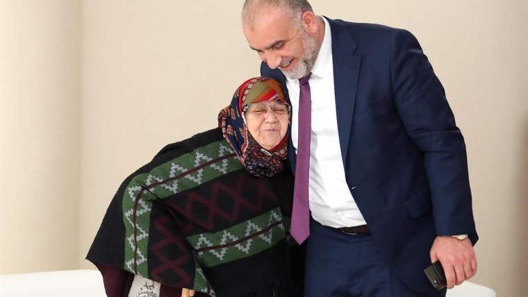 Takım elbiseli bir kişi, desenli şal takan yaşlı bir kadının yanında eğilerek poz veriyor.