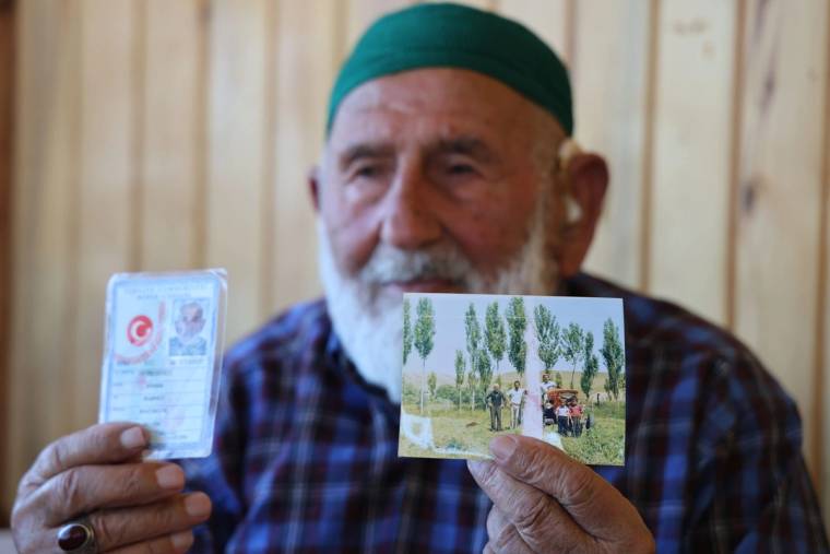 Yeşil takkeli, beyaz sakallı yaşlı bir erkek, iki elinde kimlik kartı ve bir fotoğraf tutuyor.