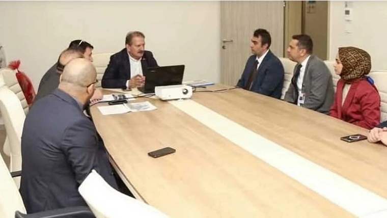 Toplantı odasında uzun masa çevresinde oturan altı kişi, projeksiyon cihazı ve belgeler görülüyor.