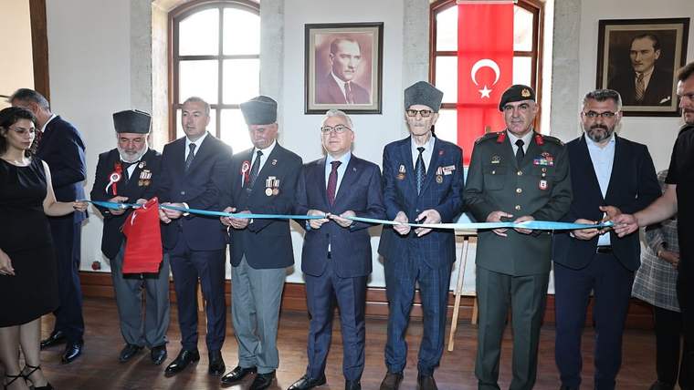 Bir salonda üniformalı kişiler ve siviller, Türk bayrağı önünde kurdele kesme törenine katılıyor.