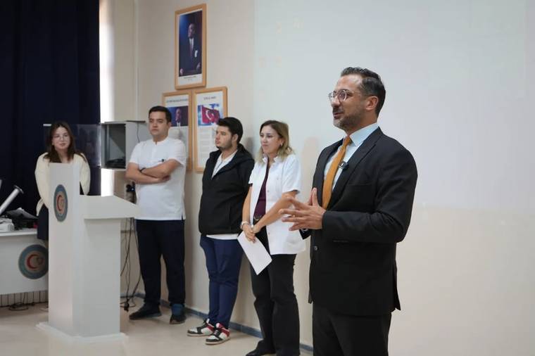 Kravatlı bir kişi konuşurken, kürsü yanında duran dört kişi ve duvardaki portreler görülüyor.