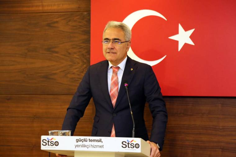 Kürsüde konuşan gözlüklü bir kişi, arkasındaki Türk bayrağı önünde duruyor; kürsüde STSO yazıyor.