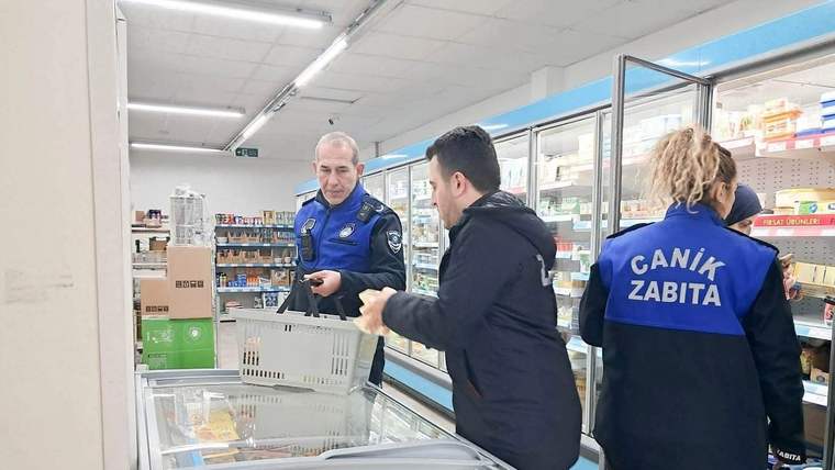 Market reyonunda üç kişi ve üzerinde “Canik Zabıta” yazan görevli, soğutucu dolapların yanında duruyor.