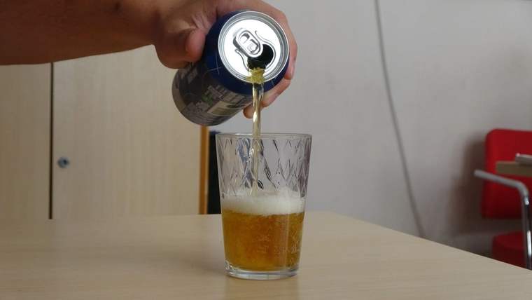 Bir el, bir kutu içeceği bir bardağa döküyor; bardakta köpüklü sarı bir sıvı var.