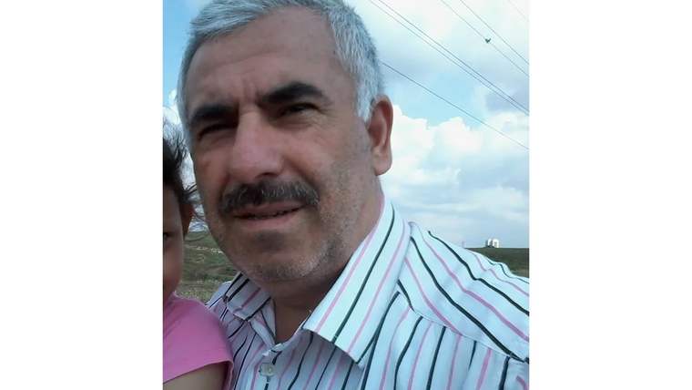 Bıyıklı bir erkek, kucağında pembe giysili bir çocukla açık havada kameraya bakıyor.