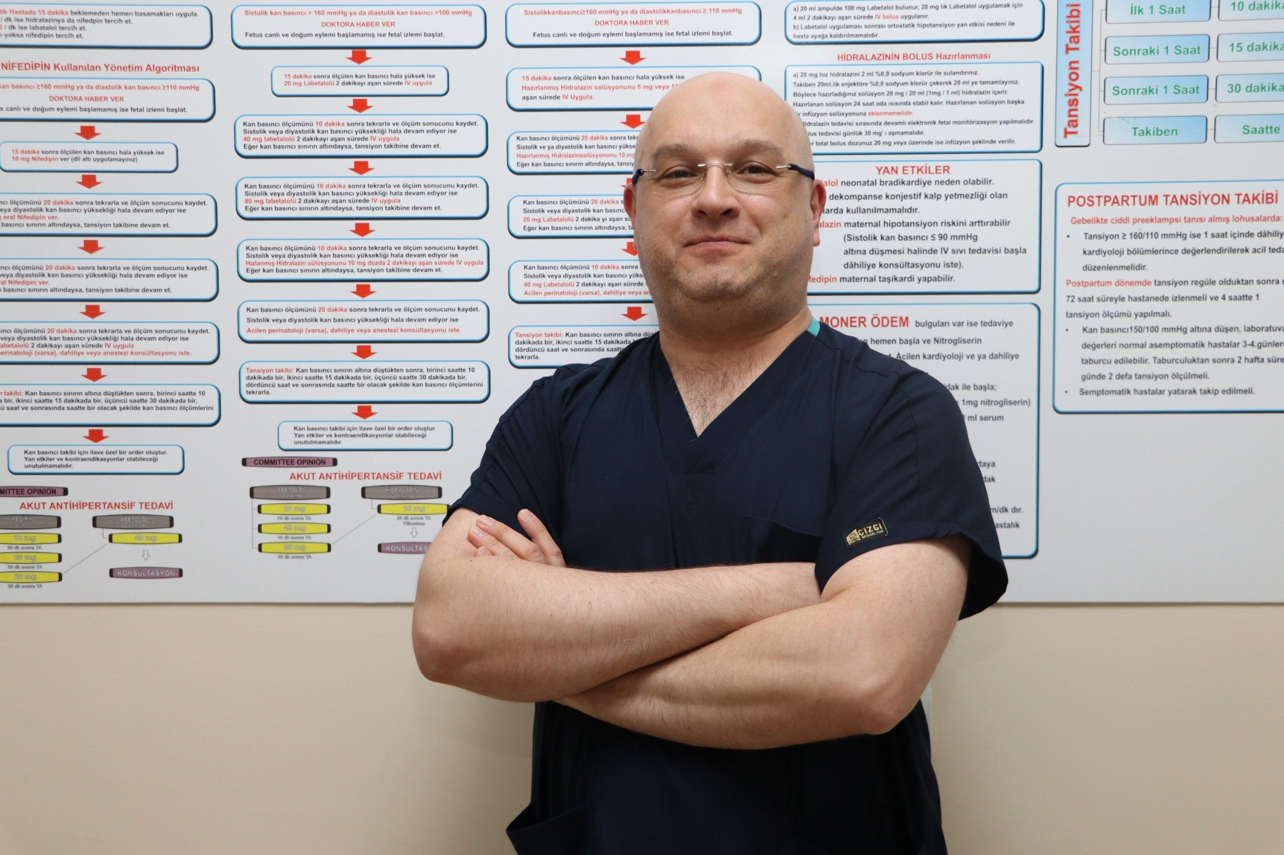 Koyu mavi kıyafet giymiş bir doktor, arkasında sağlık bilgilerini içeren bir posterle poz veriyor.