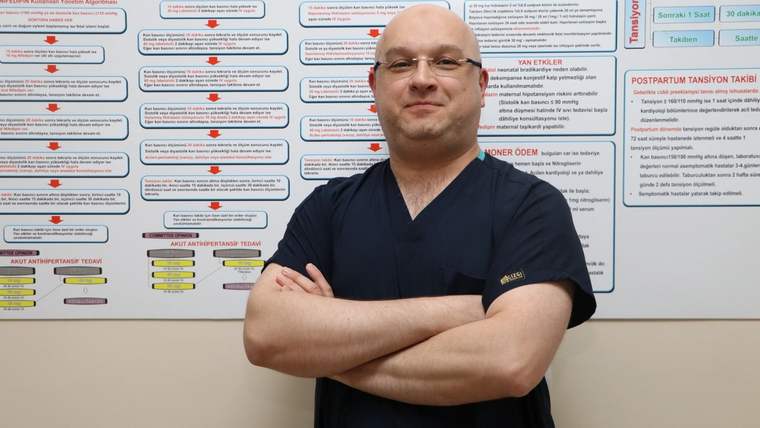 Koyu mavi kıyafet giymiş bir doktor, arkasında sağlık bilgilerini içeren bir posterle poz veriyor.