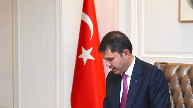 Takım elbiseli bir kişi, Türk bayrağının önünde masadaki belgeleri kalemle imzalıyor.