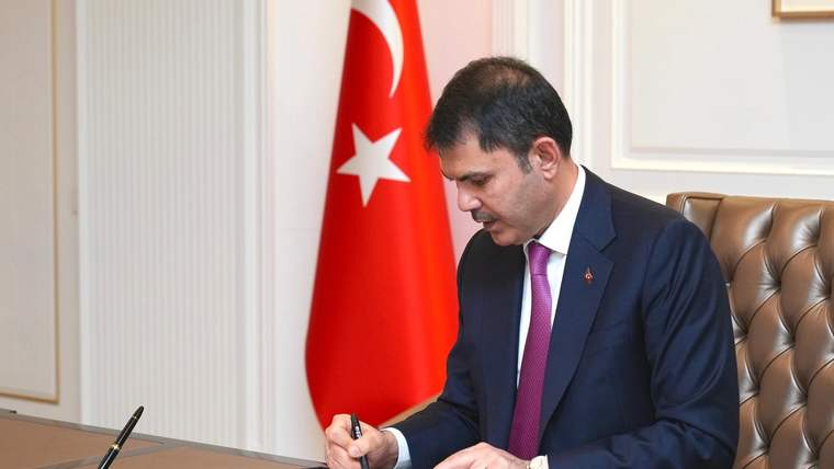 Türk bayrağı önünde takım elbiseli bir kişi, masada bulunan belgeyi kalemle imzalıyor.