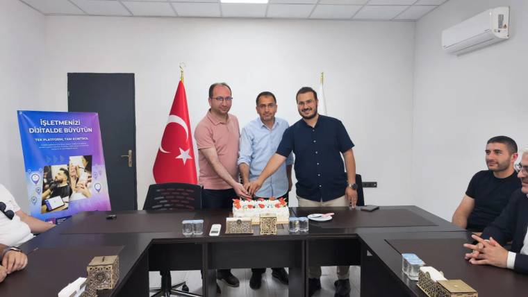 Üç kişi, masada bulunan pastayı kesiyor; arka planda Türk bayrağı ve başka bir bayrak var.