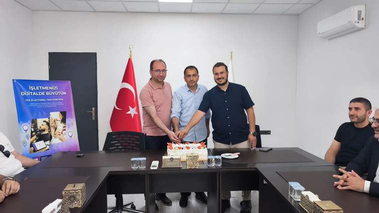 Üç kişi, masada bulunan pastayı kesiyor; arka planda Türk bayrağı ve başka bir bayrak var.