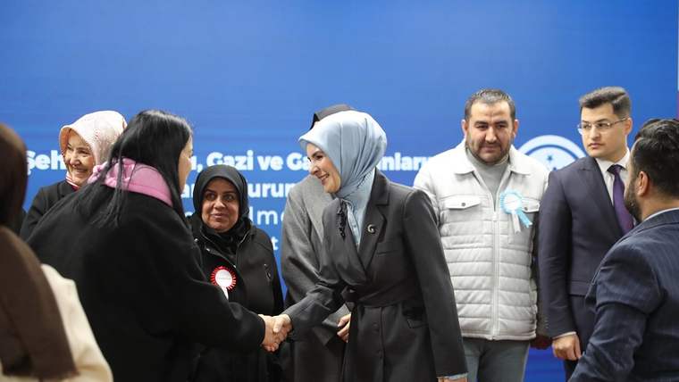 Gri başörtülü bir kadın, siyah giyimli başka bir kadınla tokalaşırken çevrelerinde insanlar duruyor.