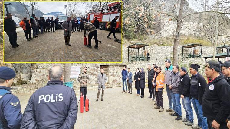 Açık alanda toplanan kişiler, kırmızı yangın tüpüyle yapılan uygulamayı izliyor; arkada itfaiye aracı görülüyor.