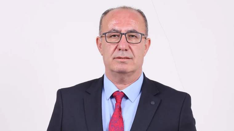 Koyu takım elbise giymiş, kravat takan bir adam, beyaz arka planda duruyor.