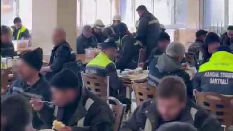 Yemekhanede çok sayıda işçi masalarda oturuyor; bazıları kaşık tutarken önlerinde tabaklar bulunuyor.
