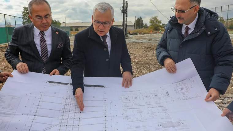 Üç kişi, açık alandaki toprak zeminde büyük mimari proje çizimlerini inceleyip üzerlerinde konuşuyor.
