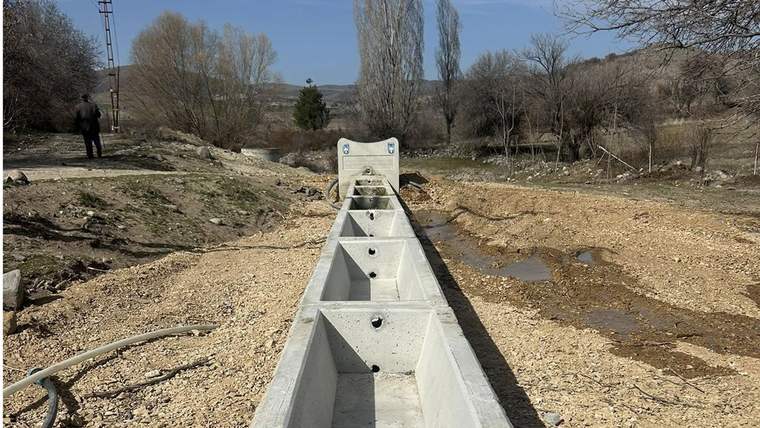 Ağaçlarla çevrili kırsal alanda, bölümlere ayrılmış uzun beton yalak ve yanında su birikintileri görülüyor.